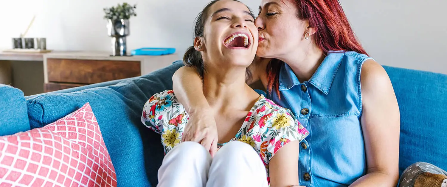 A woman with red hair, exuding warmth often found in disability care services, kisses and hugs a laughing girl on a blue couch. The girl wears a floral top and white pants, while the woman is in a denim dress. A pink patterned pillow and a small plant complete the cozy scene.