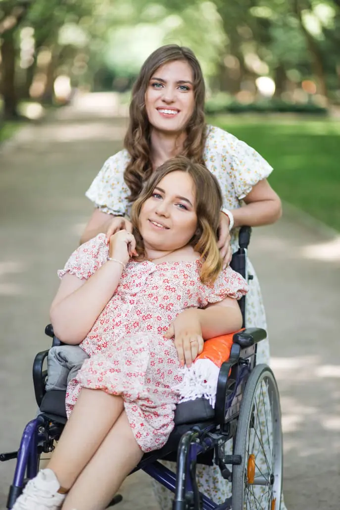 Two women are smiling outdoors on a sunny day. One is seated in a wheelchair, wearing a floral dress, embraced by sunlight and surrounded by nature's greenery. The other stands behind her, also in a floral dress, symbolizing the warmth and support of dedicated disability care services.