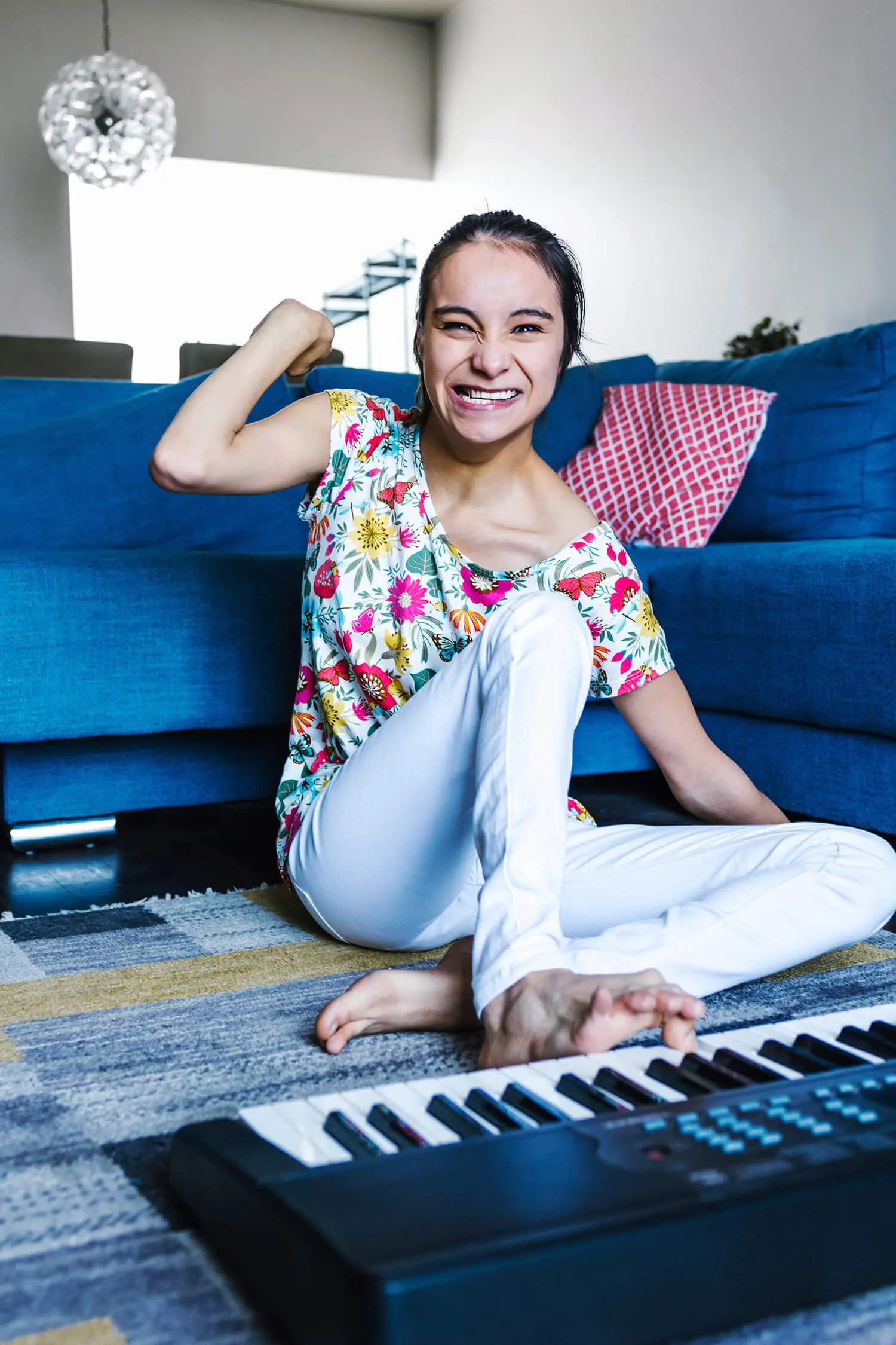 A woman in a floral top and white pants, embodying the spirit of strength often fostered by Disability Care Services, sits on the floor smiling and flexing her arm. She's near a blue couch and a keyboard, with a patterned pillow in the background.