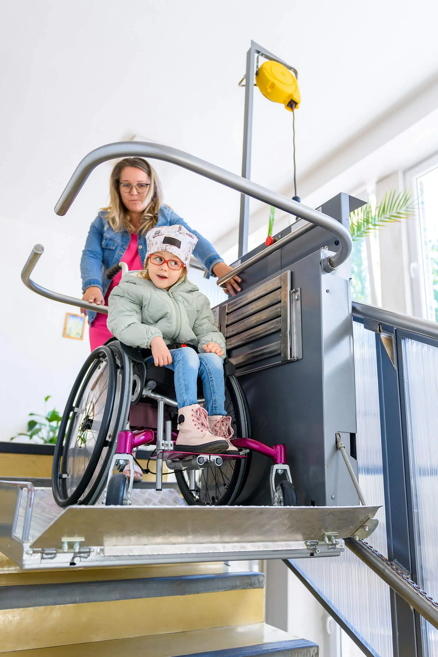 A young girl in a wheelchair is being assisted by a woman on a stairlift platform, showcasing excellent disability care services. They are going up the stairs. The girl, smiling, wears glasses, a cap, and a green jacket while the attentive woman ensures her safety.