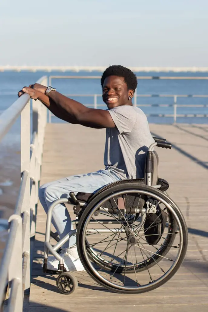 A person in a wheelchair, embodying the spirit of independence often championed by disability care services, smiles while holding onto a railing on a wooden boardwalk by the water. They're dressed in a gray t-shirt and light jeans, with wireless earbuds visible against the backdrop of blue water and clear sky.