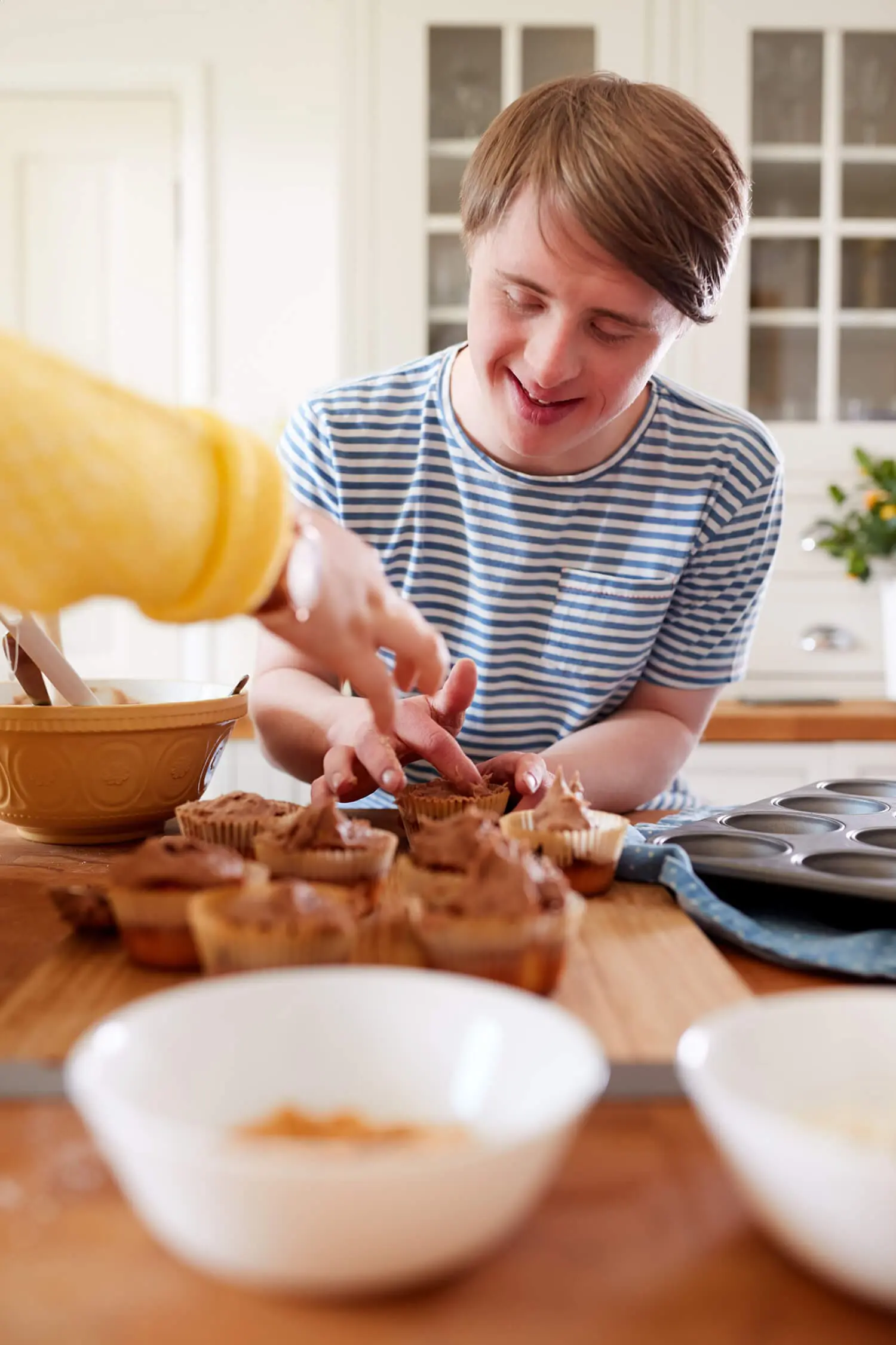 Young man in a striped shirt smiles while decorating cupcakes in a kitchen, embodying the joy of creativity that shines through disability care services. Another person reaches for a cupcake, both focused on arranging treats on a wooden countertop surrounded by baking bowls and a muffin tray.