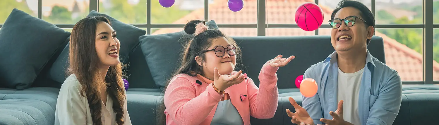 Three people sit on the floor in a sunlit living room, joyfully throwing colorful balls in the air, while several teddy bears are scattered nearby. This warm scene embodies the spirit of inclusive fun fostered by Disability Care Services.