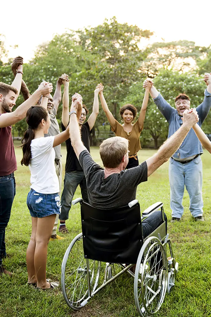 A group of diverse individuals stands in a circle outdoors, holding hands high in the air. A person in a wheelchair is part of this inclusive gathering, embodying the spirit of Disability Care Services. Everyone appears joyful, enjoying the sunny day in a grassy park area with trees in the background.