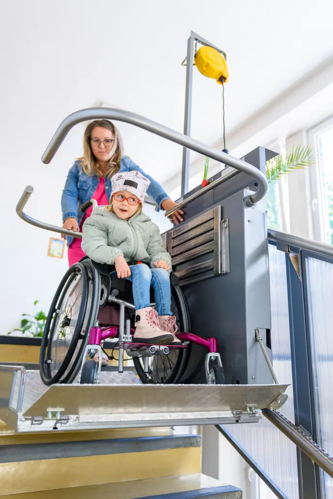 A child in a wheelchair is accompanied by a woman as they use a platform lift on the staircase, showcasing the warmth of Disability Care Services. The woman is smiling and assisting the child, who is also smiling and looking forward. The setting is indoors with natural light.