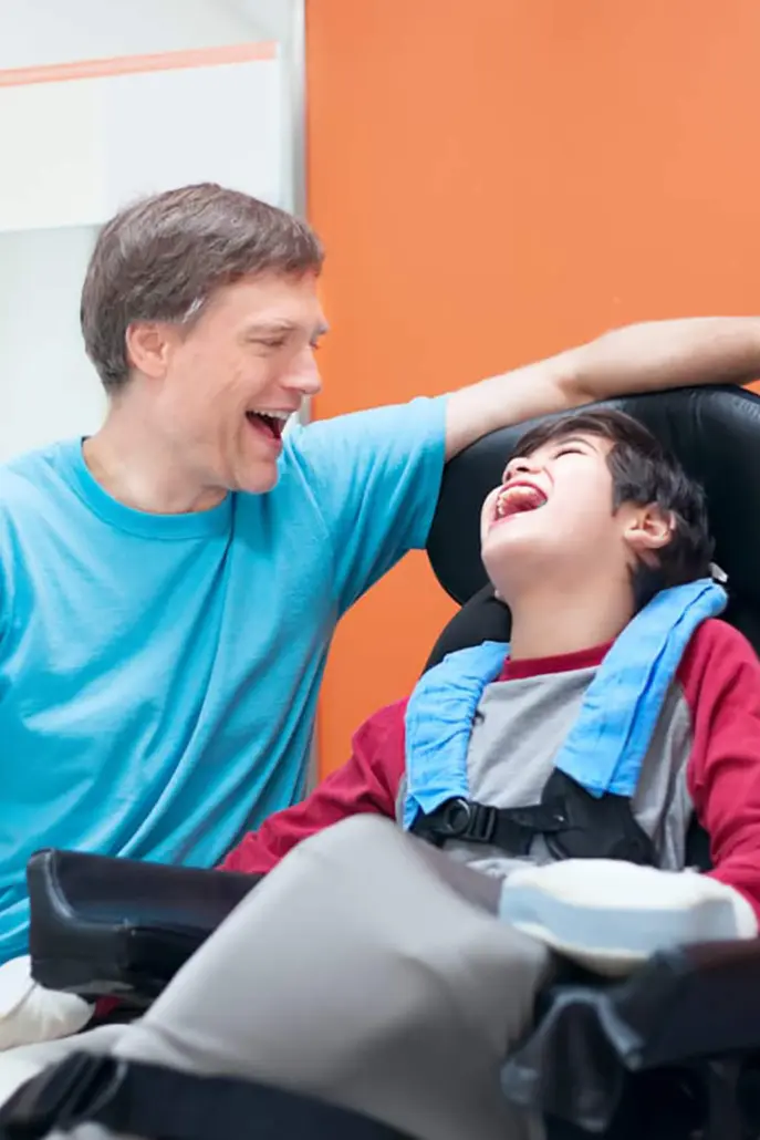 A man and a child in a wheelchair share a joyful moment, embodying the essence of disability care services. The man, in a light blue shirt, wraps his arm around the child in a maroon and gray outfit with a blue harness. Both laugh heartily against an orange and white backdrop.