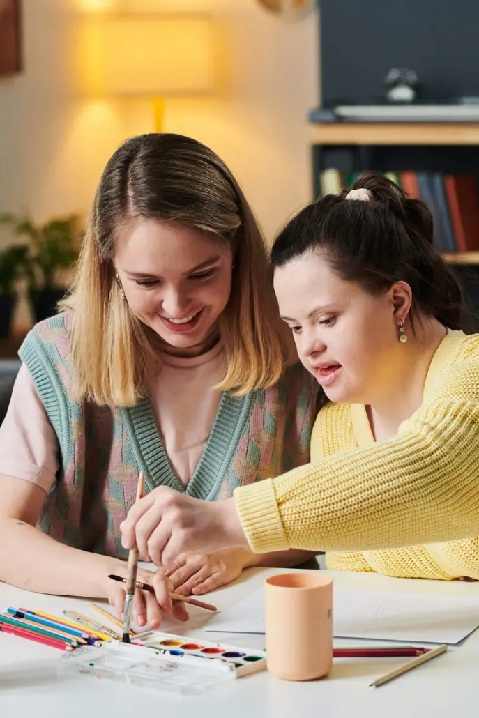 Two young women sit at a table, smiling as they paint together, illustrating the joy of creativity fostered by supportive environments like Disability Care Services. One wears a pastel-patterned vest over a pink shirt, while the other sports a yellow sweater amidst art supplies spread before them.