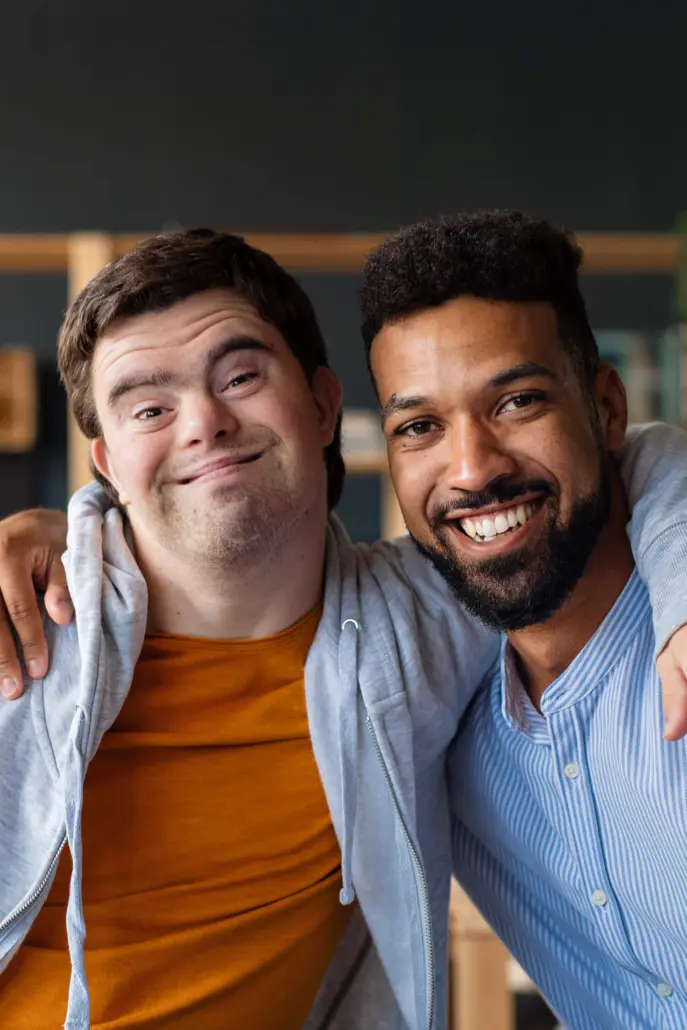 Two men smiling and embracing, one in a light gray hoodie over an orange shirt and the other in a light blue pinstriped shirt. Their warm connection radiates through the room, embodying the spirit of disability care services, with blurred background details enhancing their bond.
