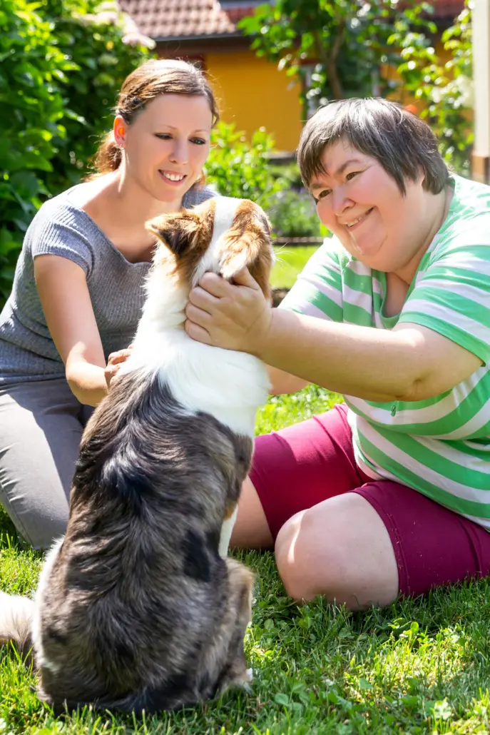 Two women sit on the grass, petting a dog. The woman on the left wears a gray shirt, while the one in a green striped shirt smiles at the dog. They enjoy a sunny garden with lush foliage and a house in the background, embodying the warmth and connection found in disability care services.