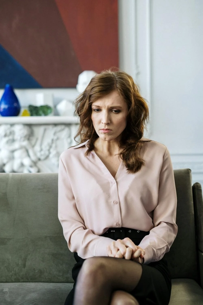 A woman with brown hair sits on a gray sofa, looking thoughtful, perhaps reflecting on her work in disability care services. She wears a light pink blouse and a black skirt. Behind her is a decorative white shelf with a blue vase, a green object, and a colorful painting on the wall.